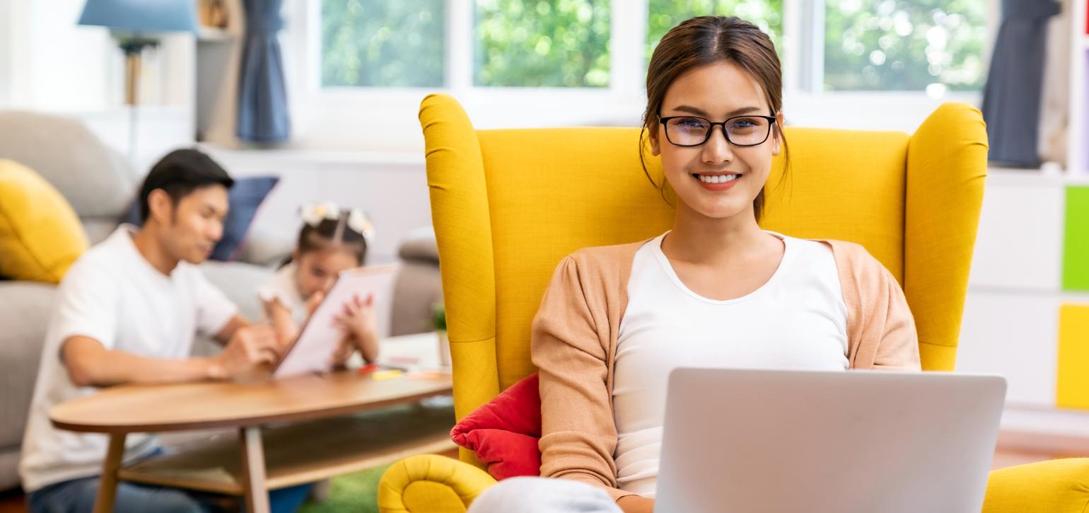 woman on laptop in yellow chair with family playing on floor behind